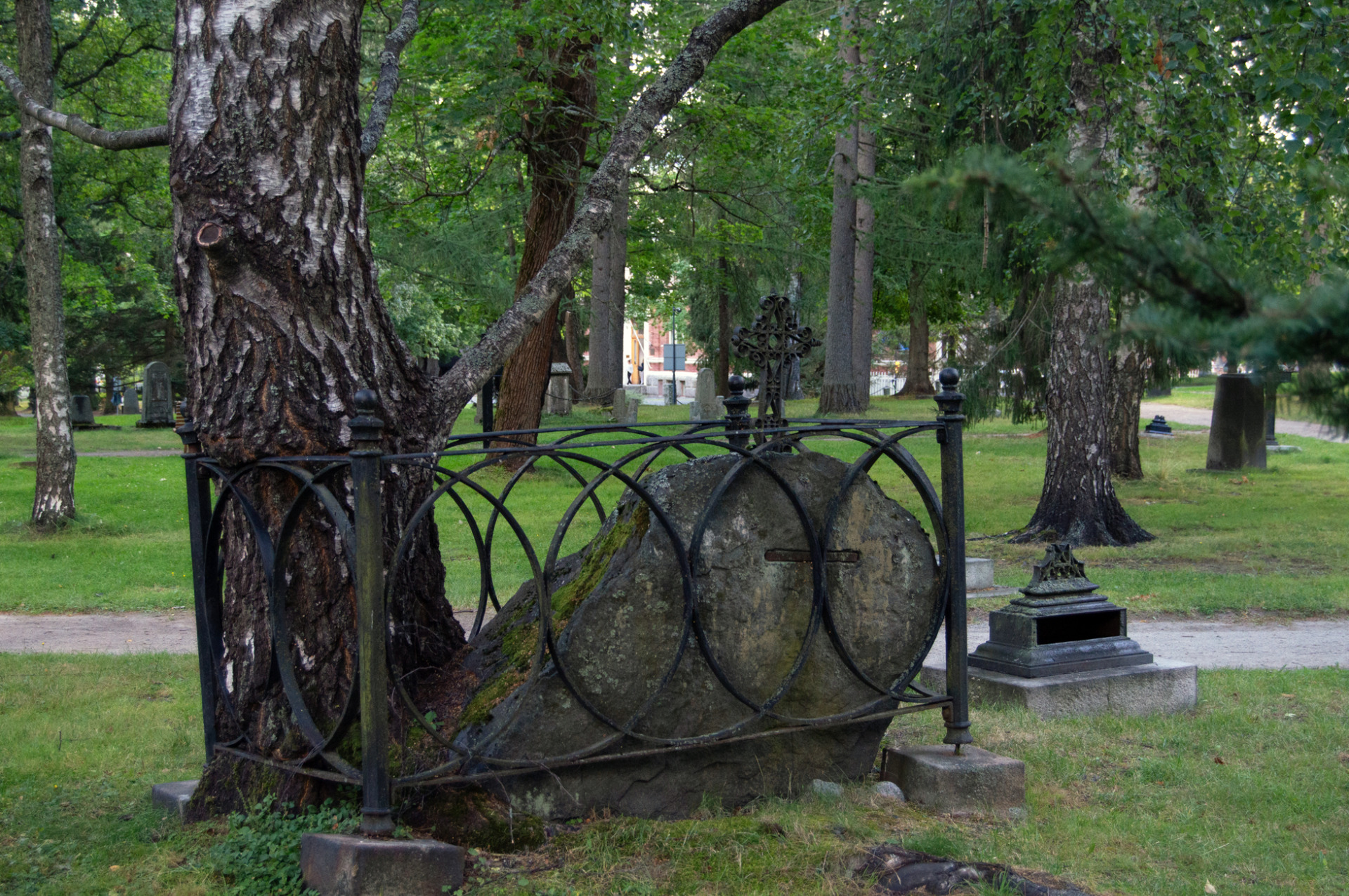 Vanha hautakivi ja rautainen seppelekehikko puiden keskellä, vihreä puisto ja muita muistomerkkejä taustalla.