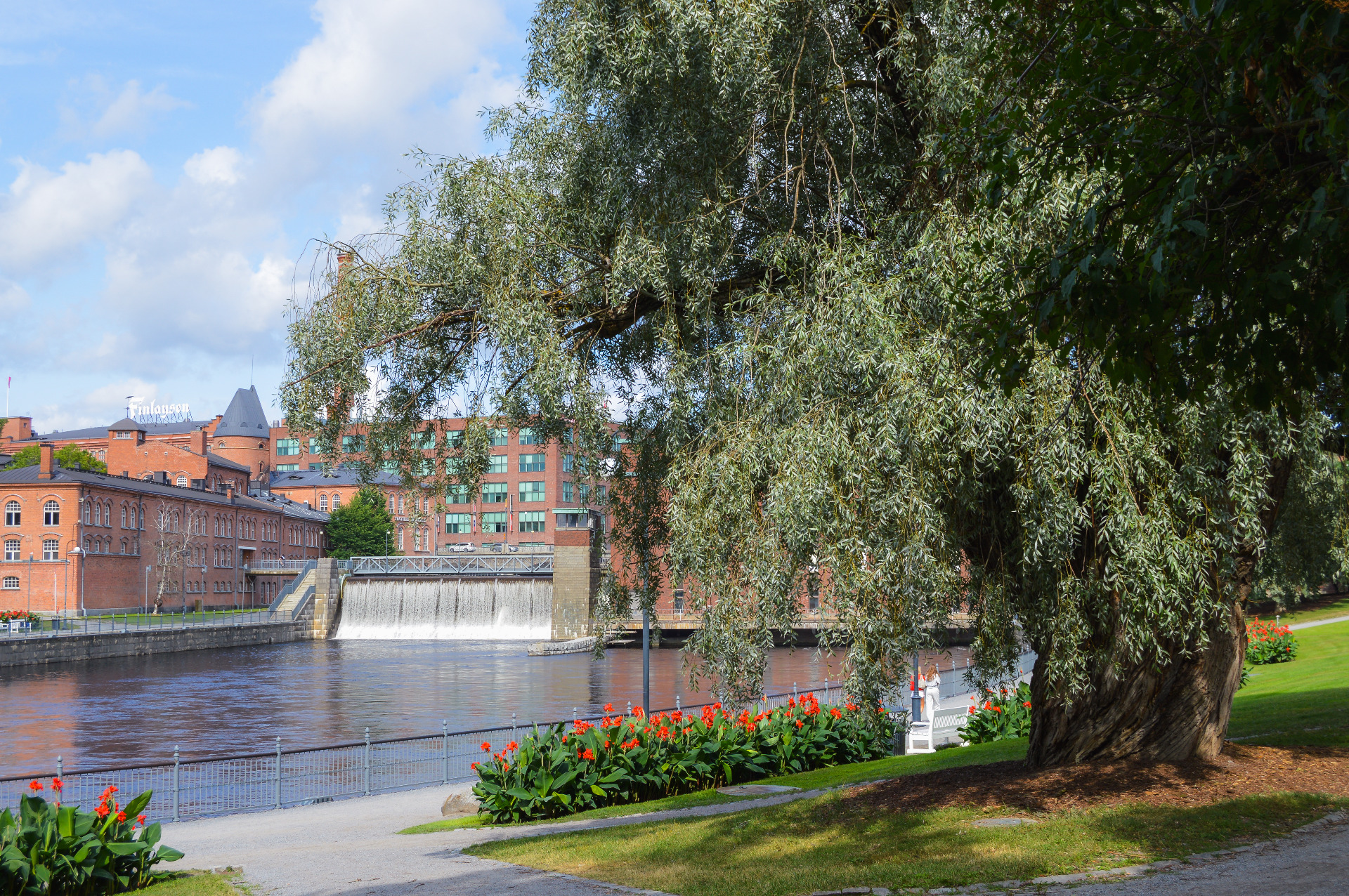 Tammerkoski, punatiilisiä rakennuksia taustalla, kukkapenkkejä ja suuri puu etualalla.