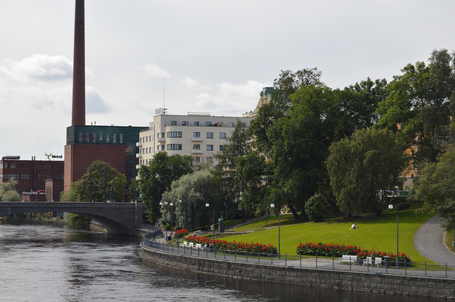 Tehdasrakennus ja korkea savupiippu kosken rannalla, puistoa, kukkapenkkejä ja kävelytie pilvisellä säällä.