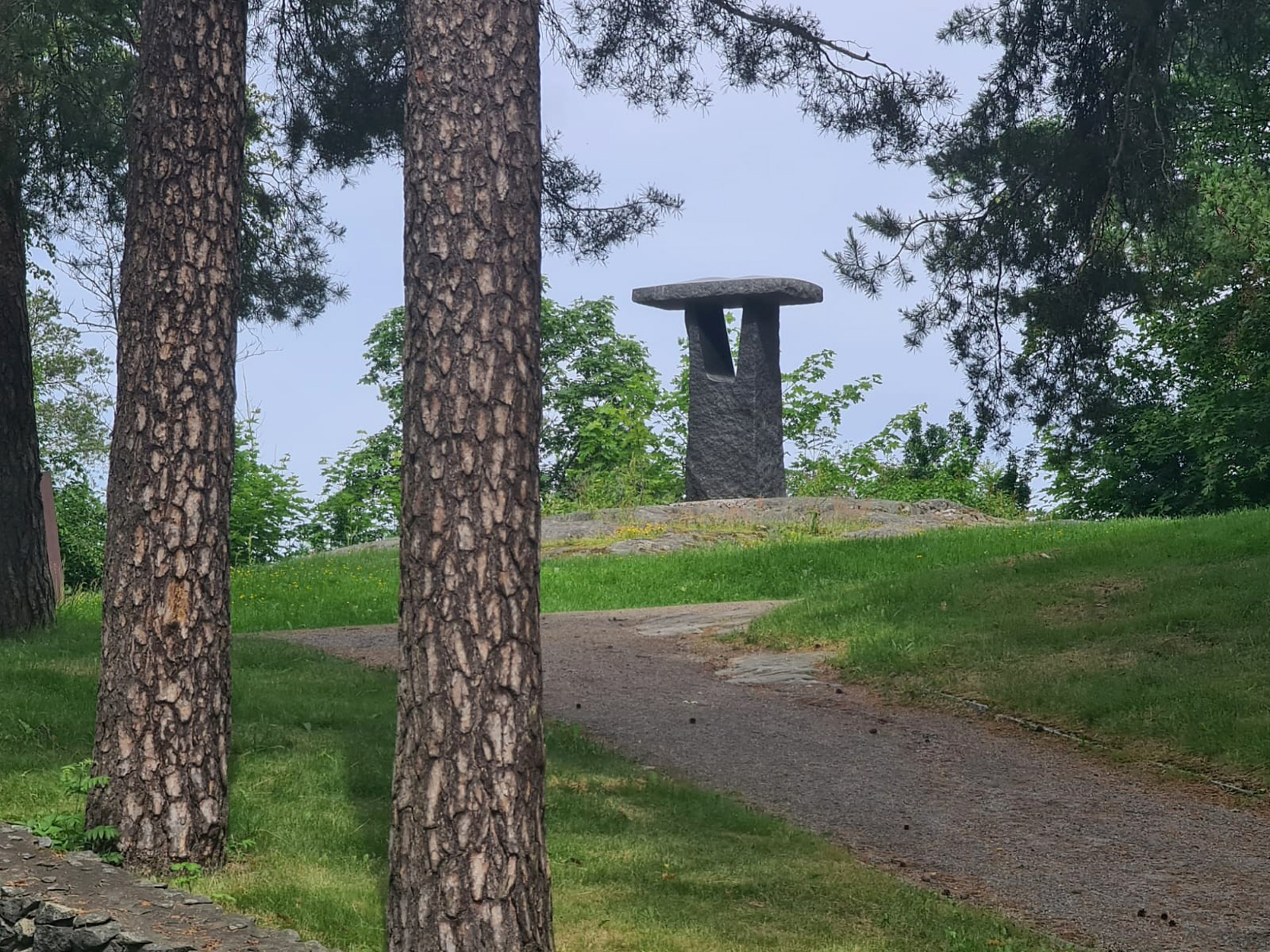 Puistopolku ja vihreä nurmikko, taustalla kivinen muistomerkki, puiden rungot etualalla.